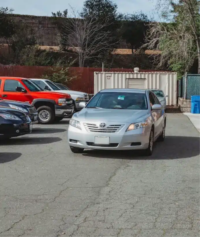 A silver sedan drives through a parking lot with other parked cars on both sides, trees in the background, and a shipping container behind the vehicles.