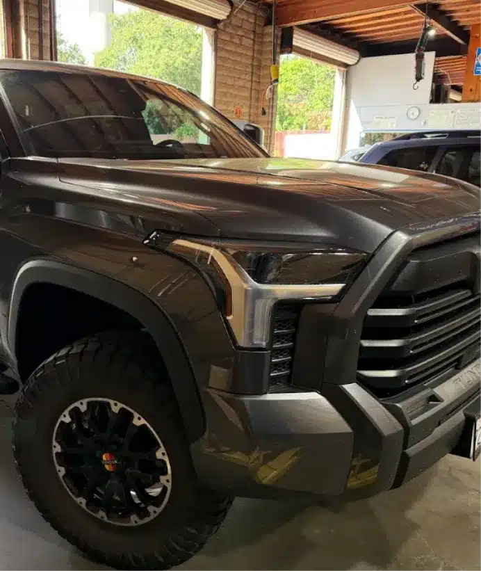 Fleet maintenance, auto repair in Walnut Creek, CA at N Service Auto Repair and Service. A close-up of the front left side of a dark gray pickup truck, showing the headlight, grille, and rugged tire, parked inside a garage with large windows and wooden beams.