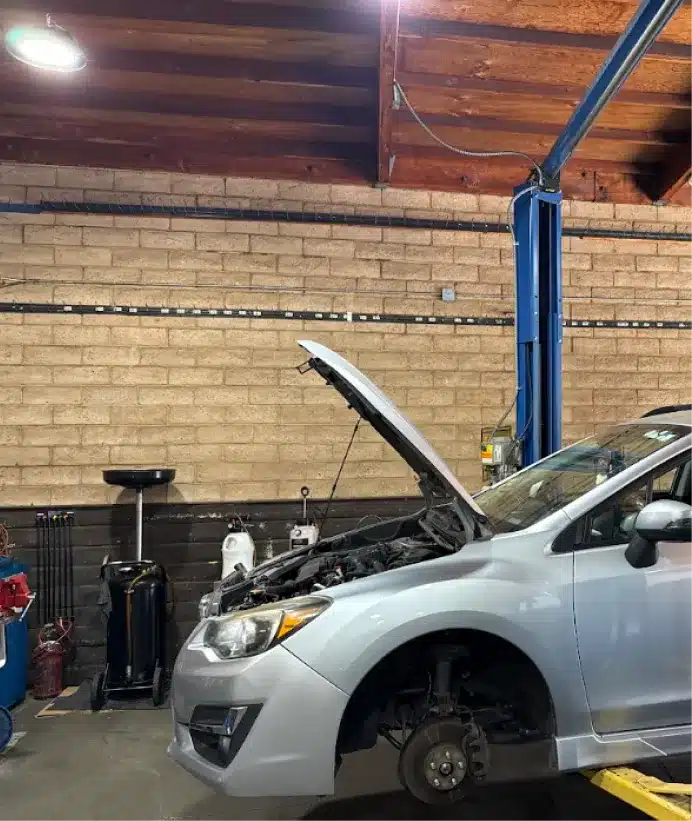 Preventative maintenance and oil change, auto repair in Walnut Creek, CA at N Service Auto Repair and Service. A silver car with its hood open and front wheel removed is parked inside a garage with brick walls and wooden ceiling. The car appears to be undergoing maintenance or repair.