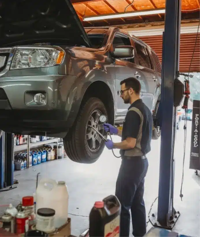 Tire services, auto repair in Walnut Creek, CA at N Service Auto Repair and Service. A mechanic wearing gloves inspects the front wheel of a gray SUV lifted on a hydraulic car lift inside an auto repair shop. Various tools, oil bottles, and car supplies are visible in the garage.