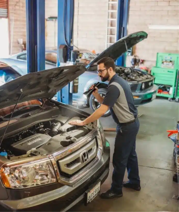 Vehicle inspections,auto repair in Walnut Creek, CA at N Service Auto Repair and Service. A mechanic inspects a Honda SUVs engine with a flashlight inside an auto repair shop. Another car with its hood open is visible in the background.