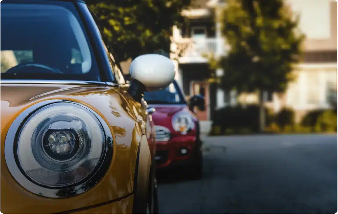 Close-up of the front of a yellow car parked on a street, with another red car parked behind it. Trees and a building are visible in the blurred background.